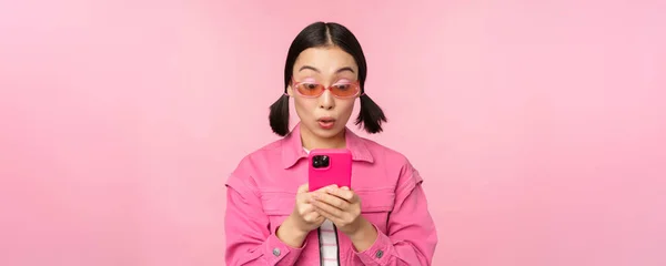 Retrato de menina asiática em óculos de sol usando smartphone. Mulher olhando para o telefone celular, navegando no aplicativo, de pé sobre fundo rosa — Fotografia de Stock