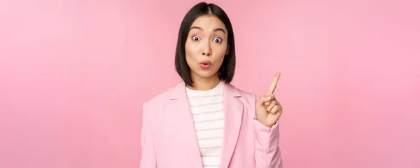 Habe eine Idee. Junge asiatische Verkäuferin, Büroleiterin hebt den Finger, deutet an, trägt Anzug, posiert vor rosa Studiohintergrund — Stockfoto
