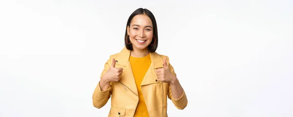 Alegre asiático mujer sonriendo complacido, mostrando pulgares arriba en aprobación, de pie en casual ropa sobre blanco fondo —  Fotos de Stock