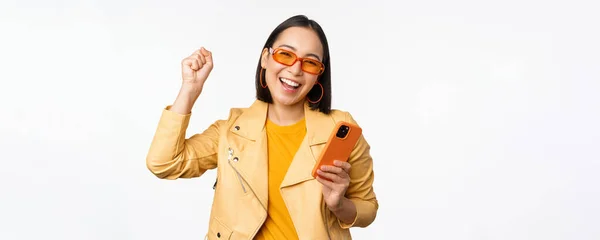 Menina coreana entusiástica em óculos de sol, segurando smartphone, celebrando e dançando, rindo feliz com o telefone celular, de pé sobre fundo branco — Fotografia de Stock