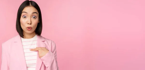 Retrato de mulher de negócios asiática reage surpreso, aponta para si mesma com descrença no rosto, posando em terno contra fundo rosa — Fotografia de Stock