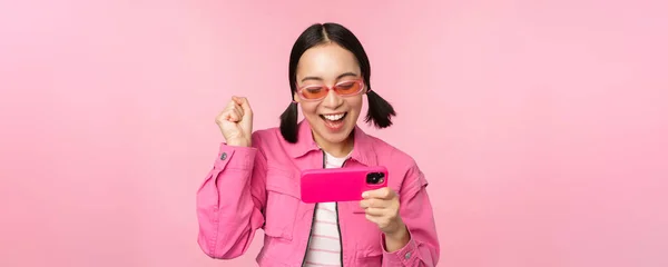 Щаслива дівчина, яка посміхається, виграє на мобільному телефоні, дивиться на горизонтальний екран смартфона і радіє, досягає мети, прославляє, стоїть над рожевим фоном. — стокове фото