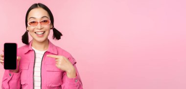 Şık giyinmiş, güneş gözlüklü, cep telefonu ekranını işaret eden akıllı telefon uygulamasını gösteren, pembe arka planda duran tutkulu Asyalı kadın.
