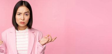 Asyalı genç bir iş kadını portresi, omuzlarını silkmiş, kafası karışmış, saf, pembe arka planda duran bir kadın.