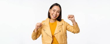 Asyalı bir kadın dans ediyor ve gülümsüyor, mutlu, zaferi kutluyor, zafer kazanıyor, beyaz arka planda duruyor.