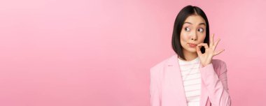 Asyalı kurumsal bir kadının portresi, ağzı mühürlü, dudakları kapalı anahtar hareketi, söz ver sır tut, pembe arka planda duran takım elbiseli.