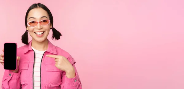 Entusiasta donna asiatica in abiti eleganti, occhiali da sole, puntare il dito contro lo schermo del telefono cellulare, mostrando applicazione smartphone, in piedi su sfondo rosa — Foto Stock