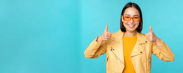 Porträtt av modern ung asiatisk kinesisk kvinna i solglasögon, bär trendiga ytterkläder, visar tummen upp och ler glad, rekommendera smth, blå bakgrund — Stockfoto