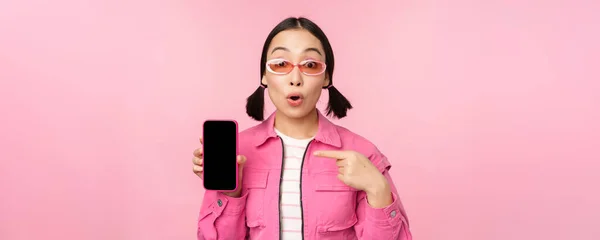 スタイリッシュな服、サングラス、携帯電話の画面で指を指す、スマートフォンのアプリケーションを示す、ピンクの背景の上に立って熱狂的なアジアの女性 — ストック写真