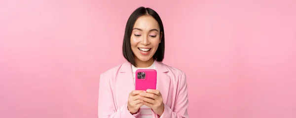 Портрет азиатской девушки в костюме со смартфоном, улыбающейся и выглядящей счастливой, стоящей на розовом фоне студии — стоковое фото