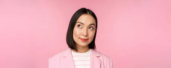 Nahaufnahme Porträt einer jungen asiatischen Geschäftsfrau, die nachdenklich lächelt und in die obere linke Ecke blickt, vor rosa Hintergrund stehend — Stockfoto