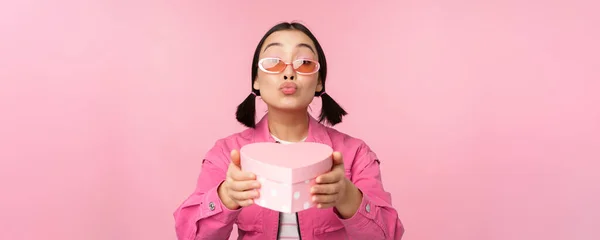 Linda chica asiática que le da regalo en forma de corazón caja, besos y sonrisas, concepto de fiesta y celebración, de pie sobre fondo rosa — Foto de Stock