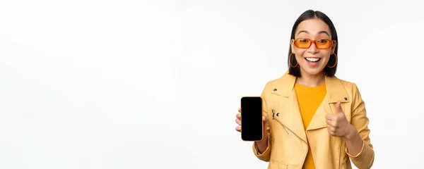 Piękna Koreanka, Azjatka w okularach przeciwsłonecznych, pokazująca interfejs aplikacji smartfona, kciuki do góry, polecająca aplikację na telefon komórkowy, białe tło — Zdjęcie stockowe