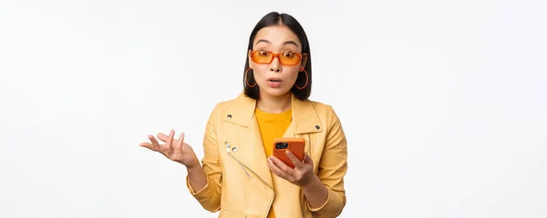 Retrato de menina coreana em óculos de sol, segurando smartphone, olhando confuso e encolhendo os ombros, de pé sobre fundo branco — Fotografia de Stock