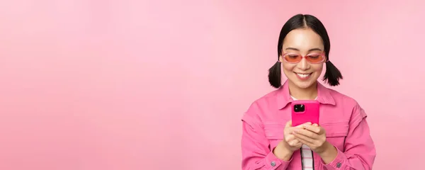 Porträtt av asiatisk flicka i solglasögon med hjälp av smartphone. Kvinna tittar på mobiltelefon, bläddrar i appen, står över rosa bakgrund — Stockfoto