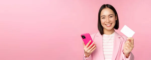Imagem de sorrir feliz asiático empresária mostrando cartão de crédito, pagando on-line no aplicativo smartphone, ordem com celular phon, de pé contra fundo rosa — Fotografia de Stock