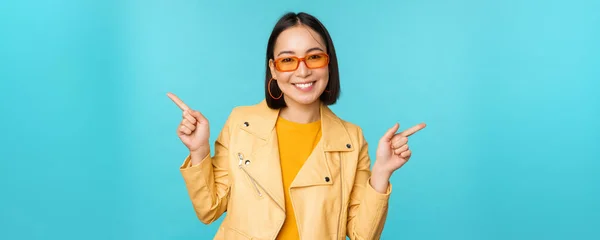 Glad snygg kinesisk flicka i solglasögon, pekar fingrarna i sidled, inbjuder till utcheckning, se butik rabatt, visar vänster och höger, står över blå bakgrund — Stockfoto