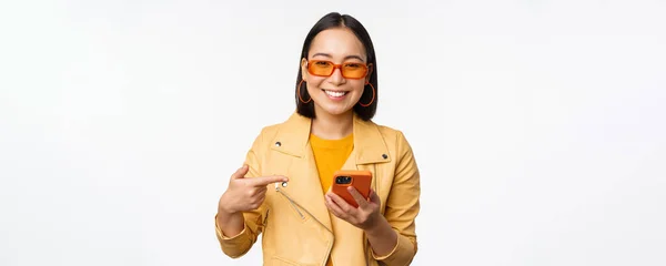 Schönes lächelndes asiatisches Mädchen mit Sonnenbrille, zeigt mit dem Finger auf Smartphone, zeigt App, speichert auf Handy, steht vor weißem Hintergrund — Stockfoto
