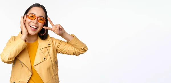 Retrato de menina moderna asiática elegante, vestindo óculos de sol e casaco amarelo, mostrando paz, gesto v-sign, de pé sobre fundo branco, rosto sorridente feliz — Fotografia de Stock