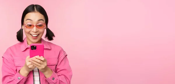 Портрет азиатской девушки в солнечных очках с помощью смартфона. Женщина смотрит на мобильный телефон, просматривает приложение, стоя на розовом фоне — стоковое фото