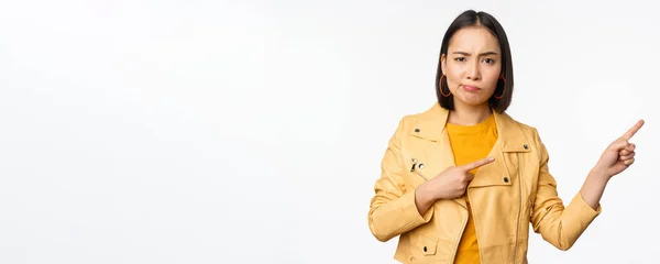 Decepcionado chica asiática frunciendo el ceño, enojado, señalando los dedos a la derecha en el anuncio, de pie sobre fondo blanco — Foto de Stock