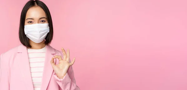 Porträtt av asiatisk affärskvinna i medicinsk ansiktsmask, visar okej tecken, bär kostym, arbetsregler under covid-19 pandemi, står över rosa bakgrund — Stockfoto