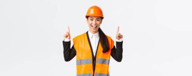İnşaat, inşaat ve endüstriyel konsept. Güvenlik kaskı ve yansıtıcı giysiler içinde mutlu gülümseyen Asyalı kadın mühendis, yeni bir nesne tanıt, satılık mülk. Mimarın işaret ettiği parmak yukarı
