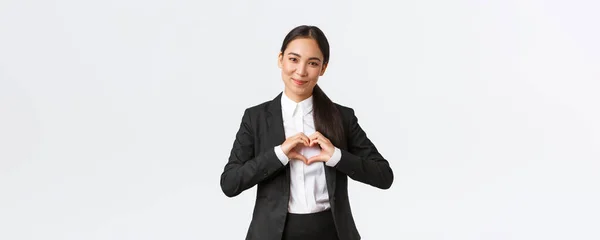Close-up van aangename Aziatische zakenvrouw, verkoopster in zwart pak zorg voor haar klanten, met hart teken en glimlach met bewondering, staande witte achtergrond zorgzaam en teder — Stockfoto