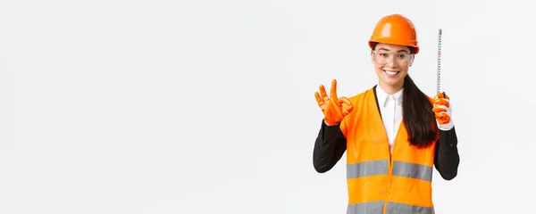 Satisfied asian female construction engineer, architect or inspector at enterprise showing okay gesture and tape measure, smiling pleased, give permission, approve measurements are valid — Stock Photo, Image