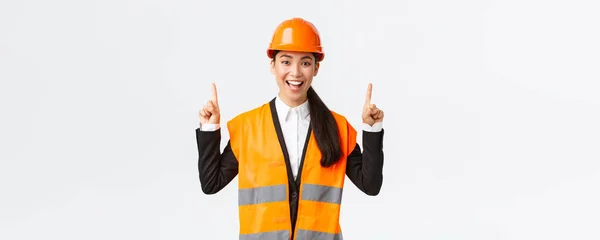 Construção, construção e conceito industrial. Feliz sorrindo engenheiro asiático feminino em capacete de segurança e roupas reflexivas, introduzir novo objeto, propriedade para venda. Arquiteto apontando dedos para cima — Fotografia de Stock