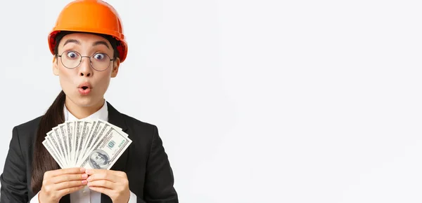 Surprised and amazed asian female engineer in safety helmet and business suit, holding money and looking astounded with big sum of income after finish construction project, white background — Stock Photo, Image
