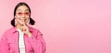Modern Koreli kız şık bahar kıyafetleri, güneş gözlükleri, sus işareti, dudaklara parmak basın, tabu jestleri, pembe arka planda dikilen