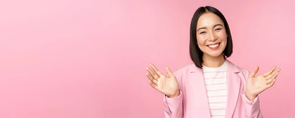 Портрет усміхненої азіатської офісної леді, бізнес-леді, що піднімає руки, махає і виглядає щасливим, позуючи в костюмі на рожевому фоні — стокове фото
