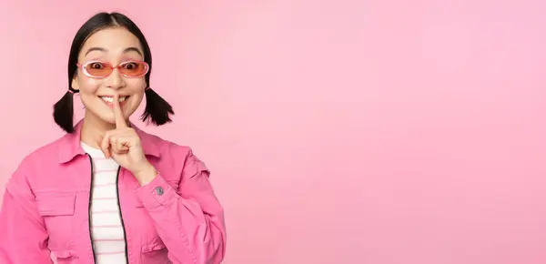 Ragazza coreana moderna in elegante abito primaverile, occhiali da sole, mostrando zittire, segno di silenzio, premere dito sulle labbra, gesto tabù, in piedi su sfondo rosa — Foto Stock