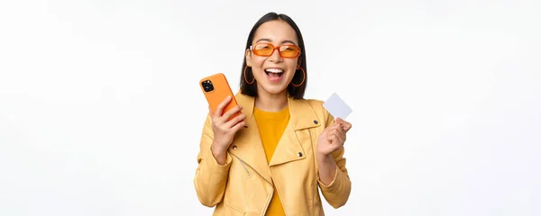 Shopping online e concetto di consegna. Felice coreano ragazza in abiti eleganti, in possesso di carta di credito e smartphone, ridere e sorridere, in piedi su sfondo bianco — Foto Stock