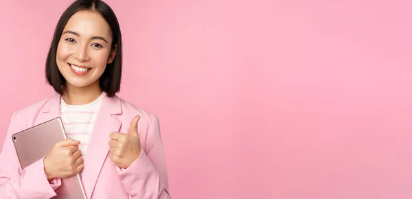 Retrato de la mujer corporativa, chica en la oficina en traje de negocios, sosteniendo tableta digital, mostrando los pulgares hacia arriba, recomendando la empresa, de pie sobre el fondo rosa —  Fotos de Stock