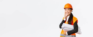 Gülümseyen mutlu Asyalı kadın mimar, güvenlik kaskı ve ceketli inşaat müdürü, bina projesinin planlarını taşıyor ve mutlu görünüyor, planı zamanında bitiriyor, beyaz arka planda duruyor.