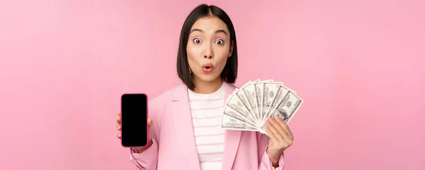 Image de coréenne femme d'entreprise réussie montrant de l'argent, dollars et écran de l'application smartphone, interface de l'application de téléphonie mobile, concept d'investissement et de finance, fond rose — Photo