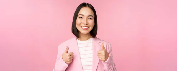 Професійна бізнес-леді, азіатська корпоративна жінка, показуючи великі пальці вгору і посміхаючись, похвали і комплімент, стоїть в костюмі на рожевому фоні — стокове фото
