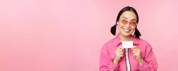 Retrato de elegante, chica asiática moderna, muestra descuento, tarjeta de crédito y mirada complacido, pagar sin contacto, concepto de compras, de pie sobre fondo rosa — Foto de Stock