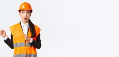 İnşaat, inşaat ve endüstriyel konsept. İlgi çekici ve şaşırmış kadın mühendis, miğfer ve yansıtıcı giysiler giyen Asyalı mimar, parmaklarını sol üst köşeyi göstererek 