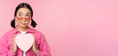 Güzel Asyalı kız mutlu gülümsüyor, kalp hediye kutusunu gösteriyor ve kameraya heyecanlı bakıyor, pembe romantik arka planda duruyor.