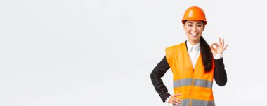 Güvenlik kaskı takmış kendine güvenen profesyonel Asyalı kadın mimar kaliteyi ve işi zamanında temin eder, iyi bir hareket gösterir ve kararlı gülümser, iddialı durur, bir şeyi garantiler ve garantiler.