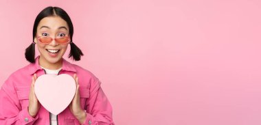 Güzel Asyalı kız mutlu gülümsüyor, kalp hediye kutusunu gösteriyor ve kameraya heyecanlı bakıyor, pembe romantik arka planda duruyor.