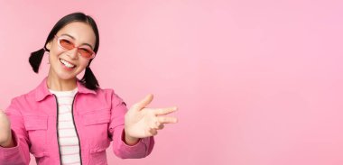 Güneş gözlüklü mutlu Koreli kız ellerini uzatıyor, gülümsüyor, dostça gülümsüyor ve pembe arka planda duruyor.