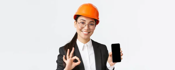 Elégedett fiatal női ázsiai mérnök, építész öltöny és biztonsági sisak mutatja a mobiltelefon képernyőn, és hogy rendben gesztus, jóváhagyja, ajánljuk alkalmazás, fehér háttér — Stock Fotó