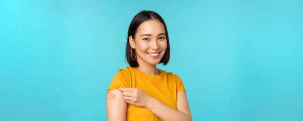 Vakcinační kampaň z covid-19. Mladá krásná, zdravá asijská žena ukazující rameno s obvazem, koncept očkování, stojící nad modrým pozadím — Stock fotografie