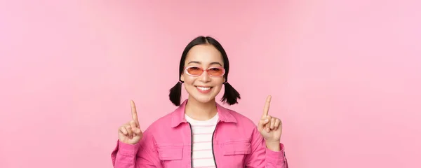 Primer plano retrato de modelo femenino coreano moderno, lleva gafas de sol, señala los dedos hacia arriba, muestra publicidad, pancarta promocional, fondo rosa —  Fotos de Stock