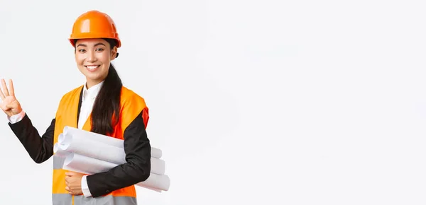 Sorrindo confiante asiático engenheiro feminino, gerente de construção em capacete de segurança, realizar plantas, mostrando quatro dedos, garantir a construção terminou a tempo, pé fundo branco otimista — Fotografia de Stock