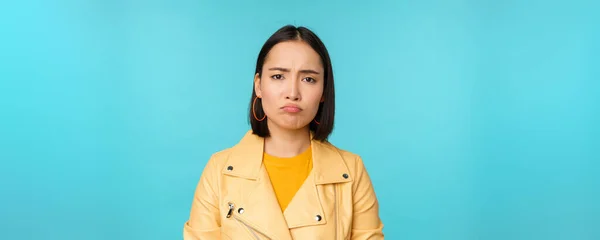 Close up of disappointed asian girl sulking, grimacing and frowning displeased, standing moody against blue background — Stock Photo, Image